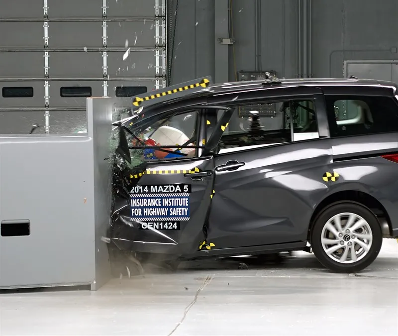 The 2014 Mazda 5 against the barrier mid-crash, with the driver's door visibly unlatched.